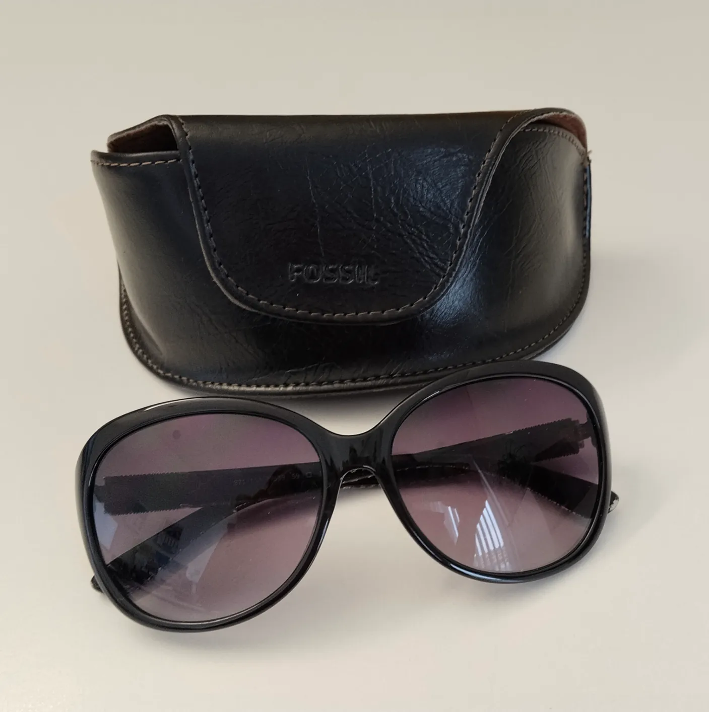Ein Paar schwarze Sonnenbrille mit passender Lederhülle. Die Sonnenbrille hat einen rechteckigen Rahmen und dunkle lila Gläser. Die Hülle ist aus schwarzem Leder mit einem weißen Logo auf der Vorderseite.