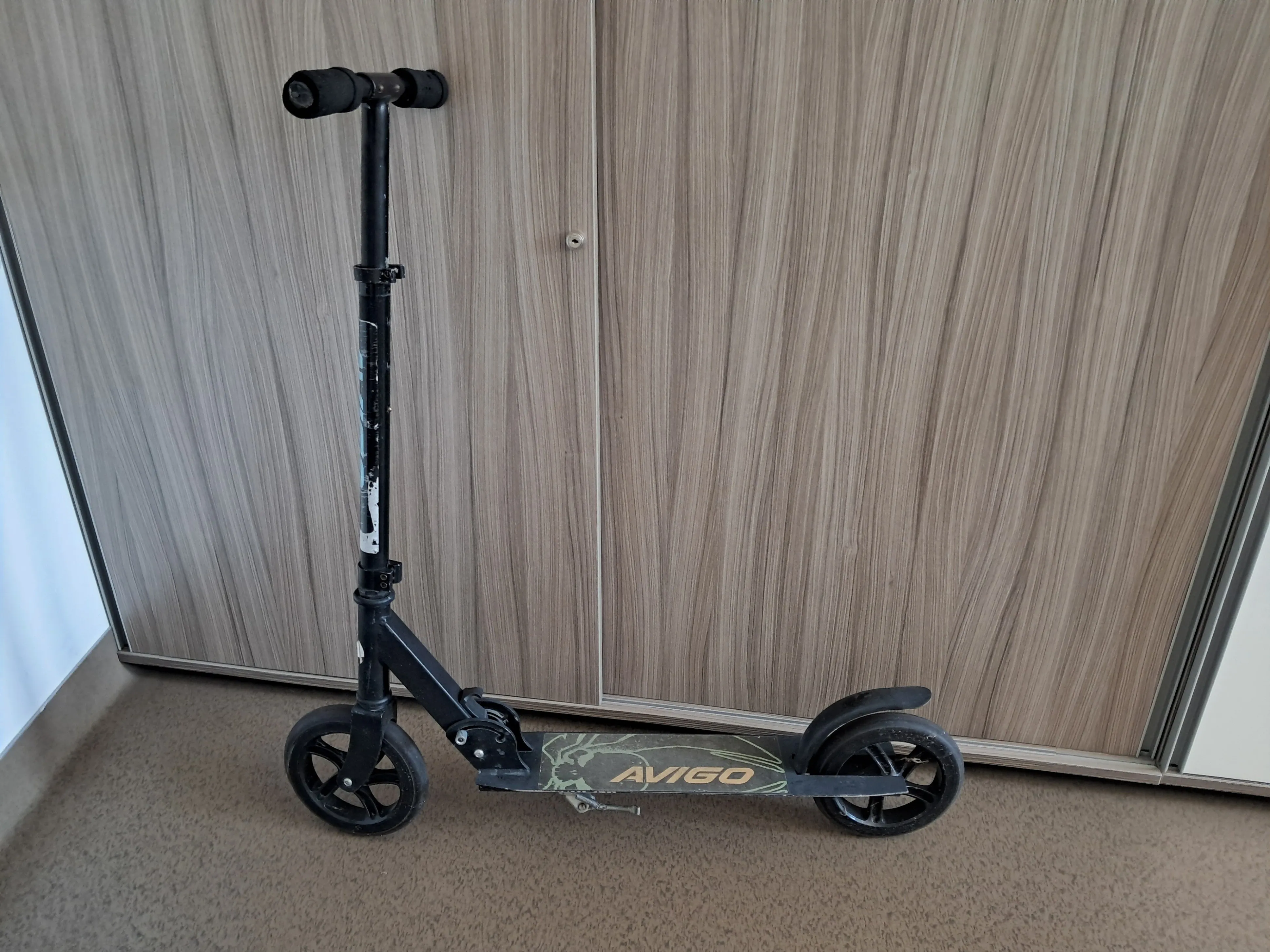 Ein schwarzer Scooter mit Tarndesign steht auf einem Holzboden vor einer Holzwand parkiert.