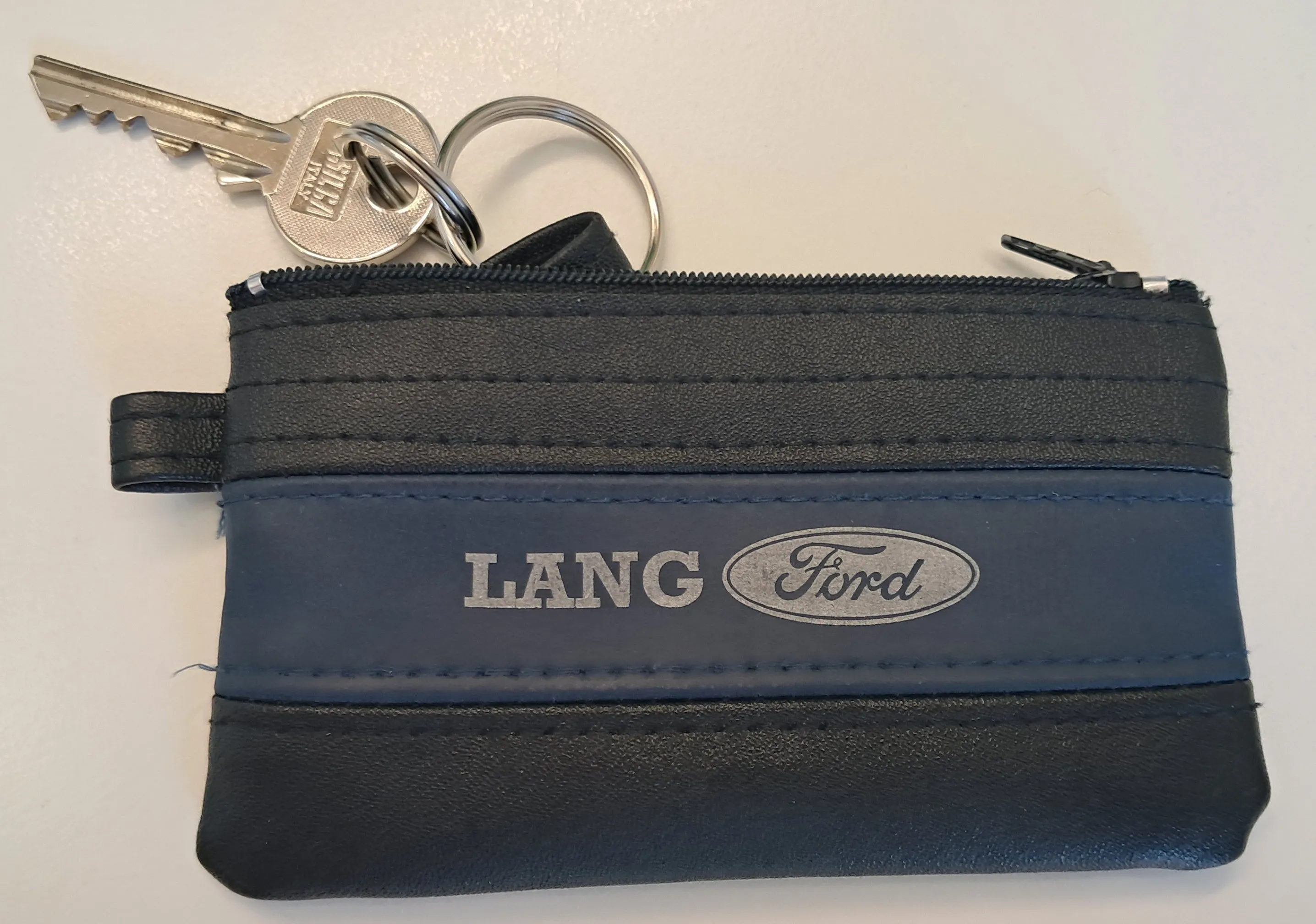 Eine blaue Ledergeldbörse mit dem Ford-Logo und dem Wort LANG darauf. Sie hat einen Reißverschluss und einen Schlüsselring mit zwei Schlüsseln.