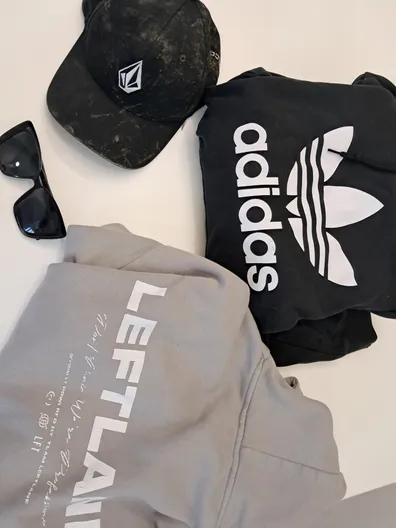 Drei Kleidungsstücke und ein Paar Sonnenbrille auf einer weißen Oberfläche. Eine schwarze Kappe, ein grauer Hoodie, ein schwarzes Adidas Sweatshirt und schwarze Sonnenbrille.