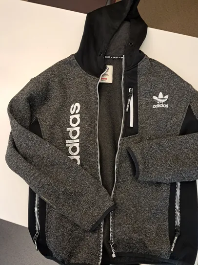 Eine graue Adidas-Hoodie-Jacke mit schwarzen Reißverschlüssen und dem Adidas-Logo auf der rechten Seite. Die Jacke hat weißen Text auf der linken Seite und ist zur Hälfte gefaltet.