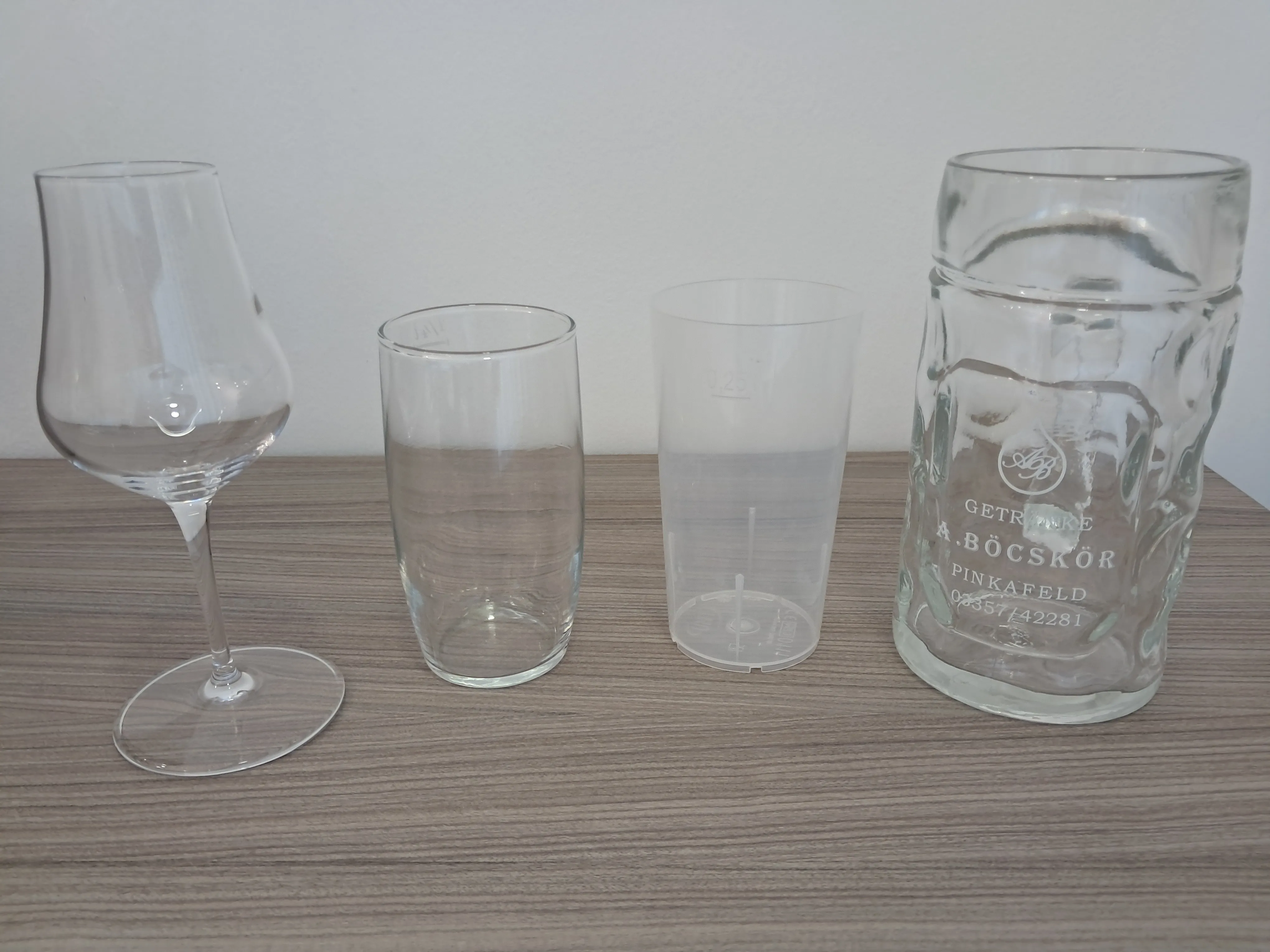 Vier leere Glasgegenstände auf einer Holzoberfläche. Von links nach rechts: ein Weinglas, ein hoher Glas, ein Plastikbecher und ein Glaskrug mit graviertem Text.