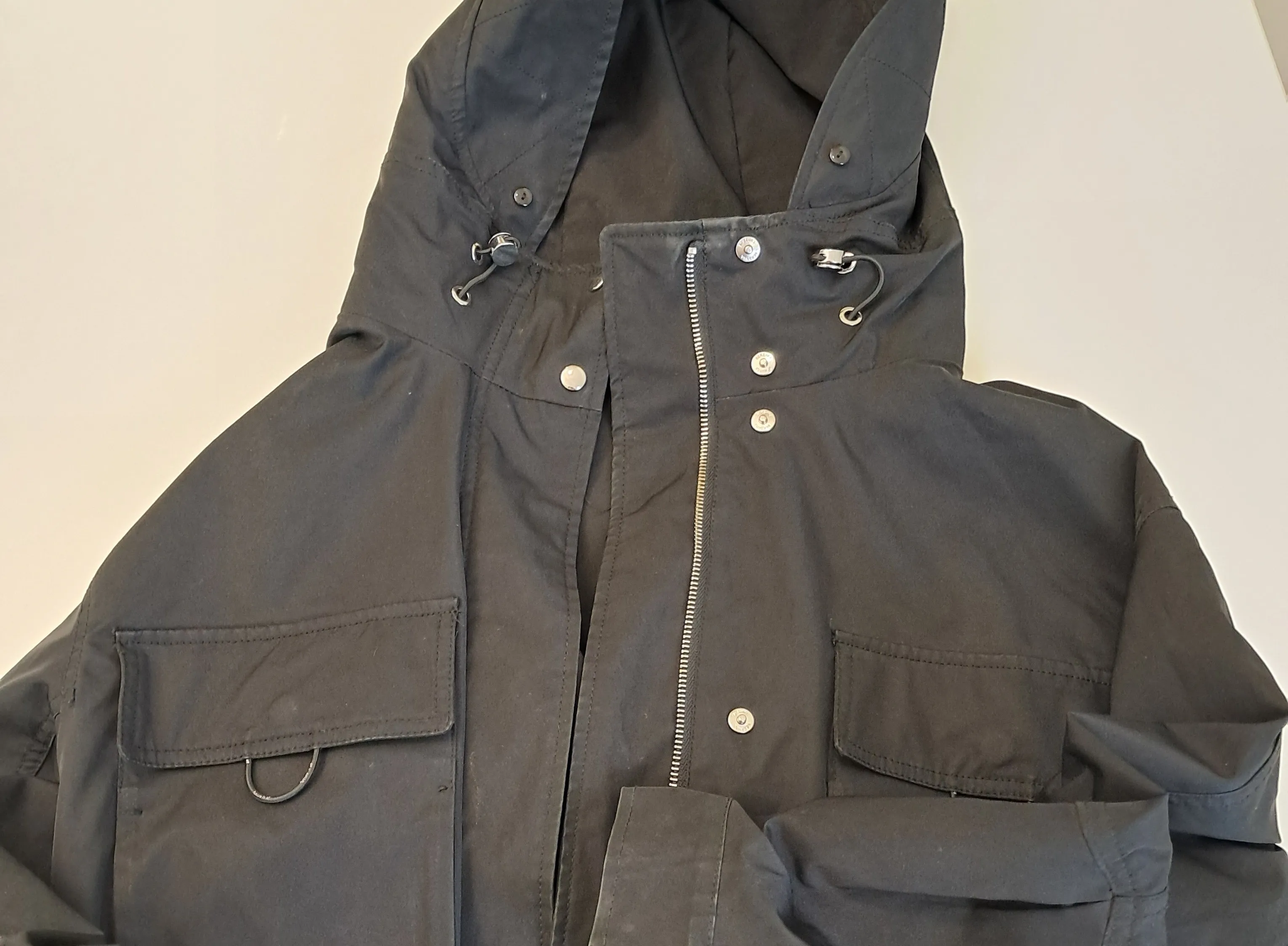 Eine schwarze Jacke mit Kapuze und Reißverschluss, mit mehreren Taschen und Knöpfen auf der Vorderseite. Die Jacke scheint aus einem robusten Material zu sein.