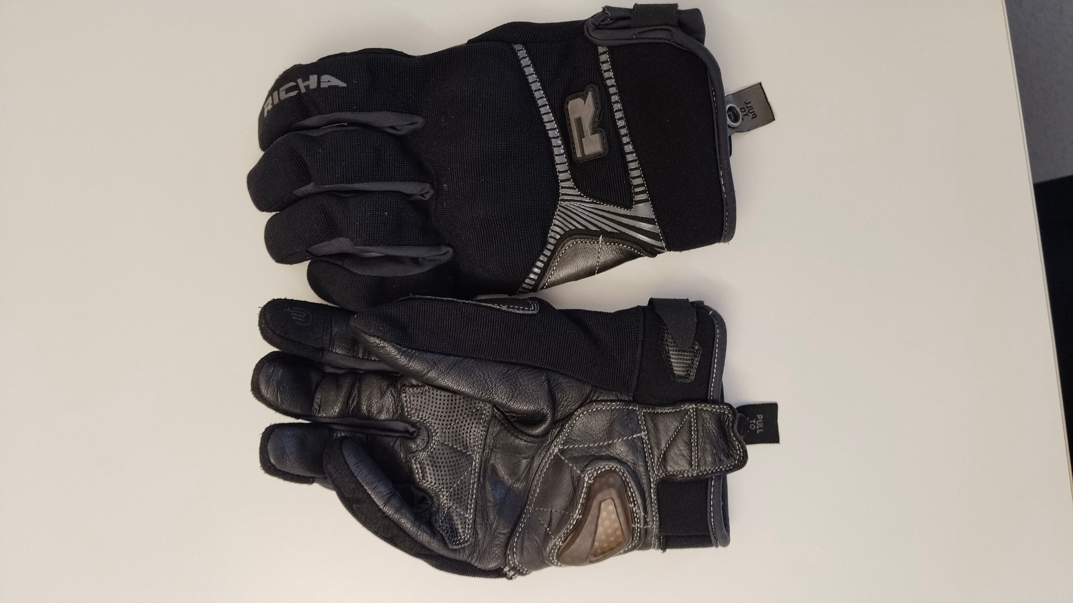 Ein Paar schwarze und graue Handschuhe mit dem Buchstaben R auf der Rückseite. Die Handschuhe haben einen Reißverschluss und einen Anhänger an der Seite.