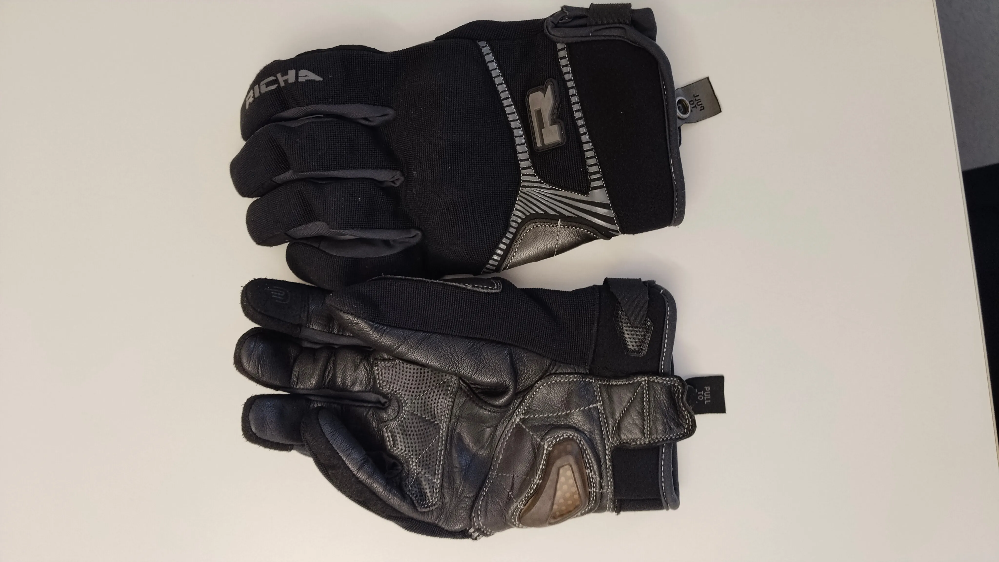 Ein Paar schwarze und graue Handschuhe mit dem Buchstaben R auf der Rückseite. Die Handschuhe haben einen Reißverschluss und einen Anhänger an der Seite.