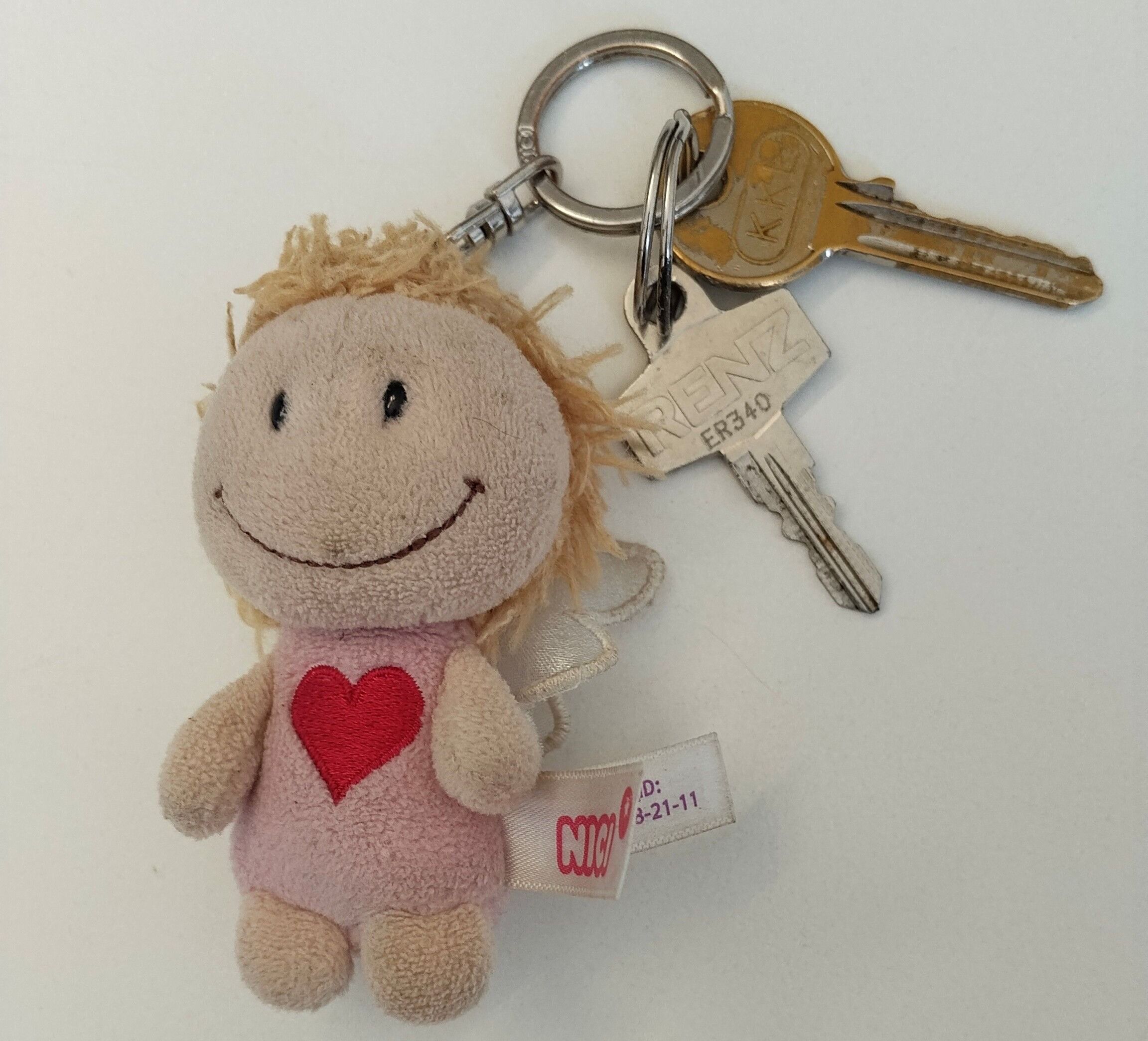 Ein kleiner Plüsch-Schlüsselanhänger mit einer lächelnden Puppe mit einem Herz auf der Brust, befestigt an einem Schlüsselring mit zwei Schlüsseln, die mit 'RENZ' und 'ER340' beschriftet sind.