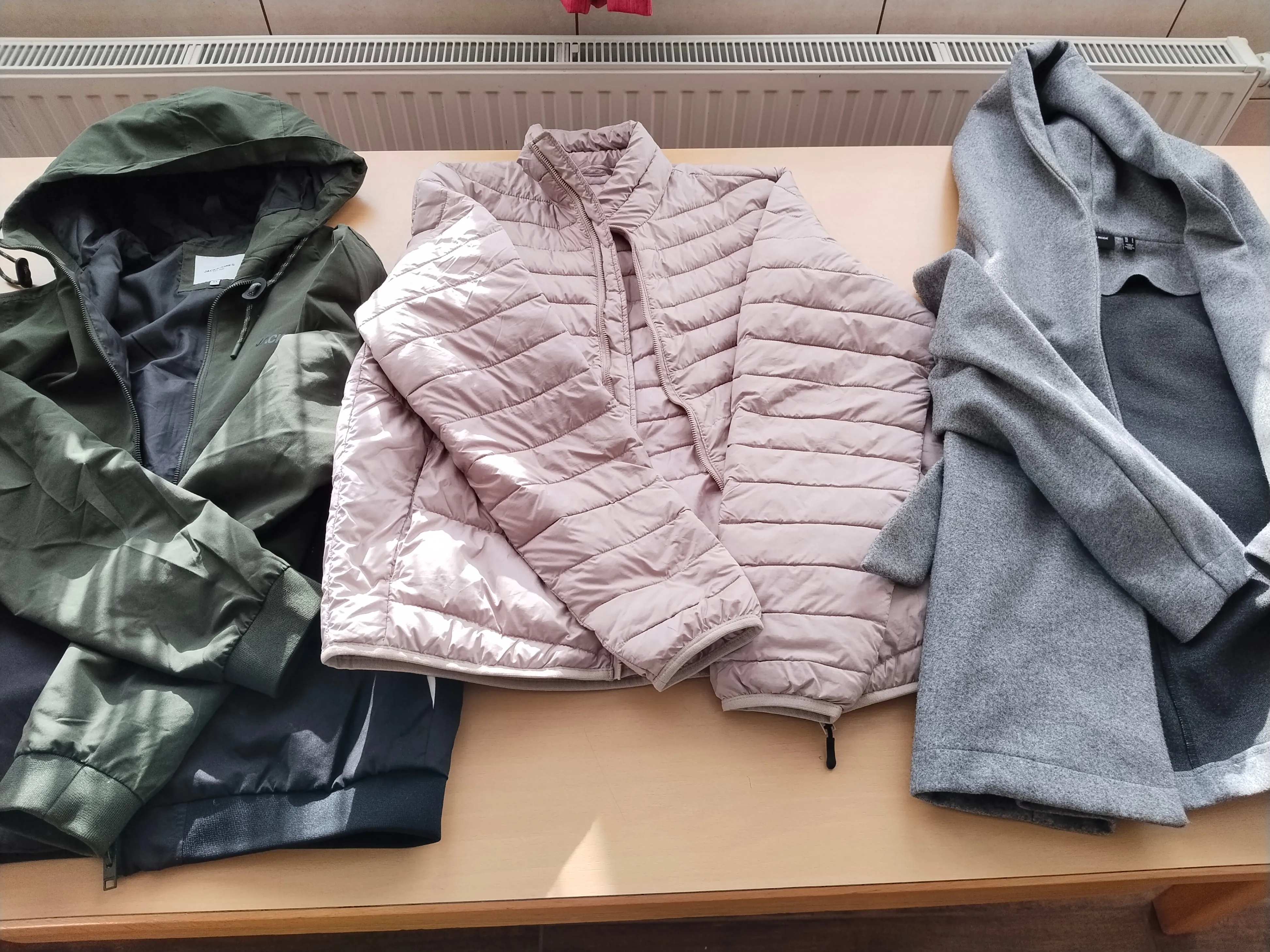 Vier Jacken sind auf einem Holztisch ausgebreitet. Eine rosa Pufferjacke ist prominent ausgestellt, neben einer grauen Kapuzenjacke, einer grünen Kapuzenjacke und einer blauen Jacke. Hinter den Jacken ist ein weißer Heizkörper an der Wand montiert.