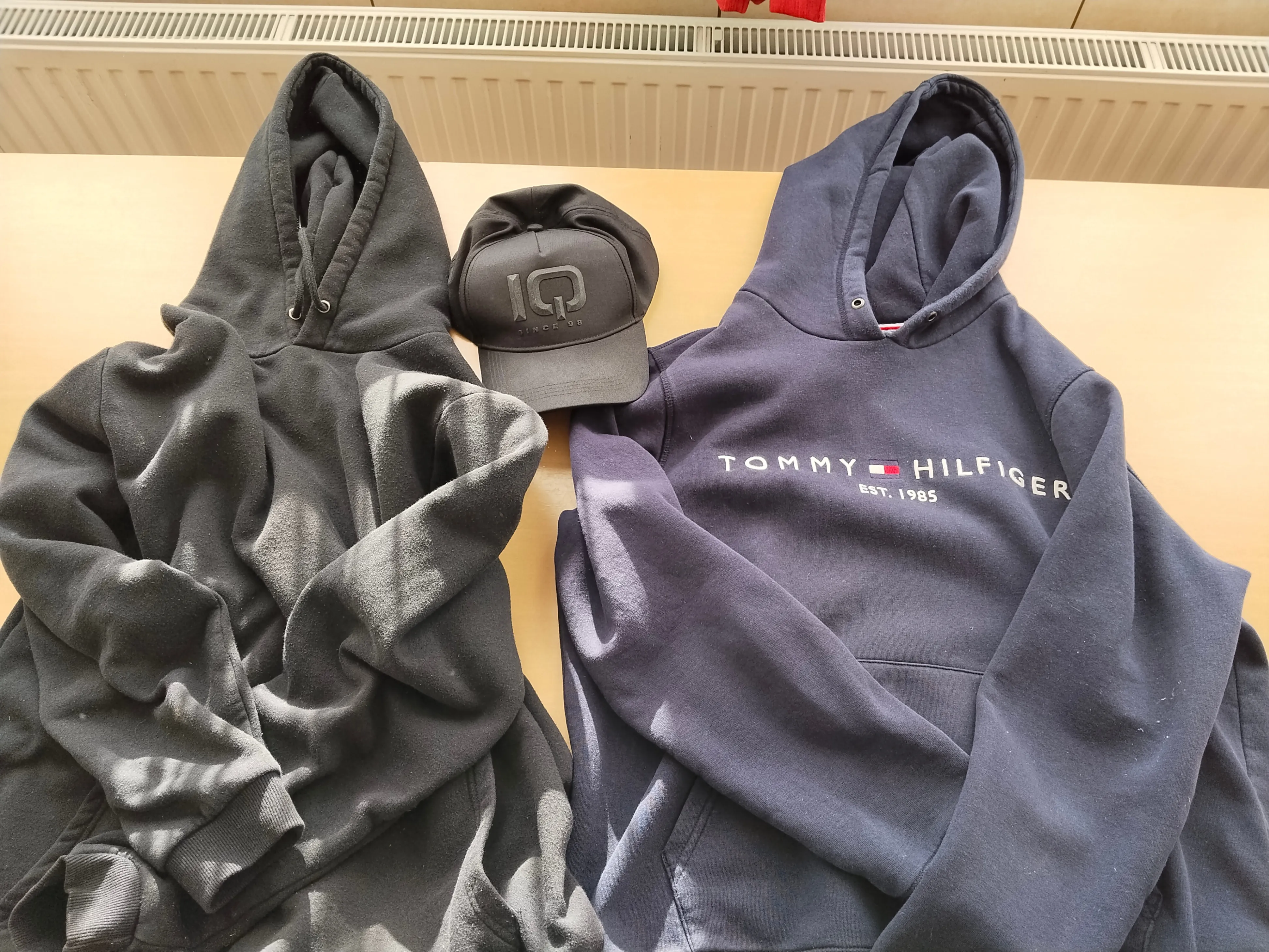 Drei graue Kleidungsstücke: zwei Hoodies und eine Kappe mit den Buchstaben IQ, alle auf einer Holzoberfläche platziert.