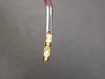 Ein vergoldeter Ring mit Diamantmuster hängt an einer silbernen Pinzette. Der Hintergrund ist eine feste graue Farbe.