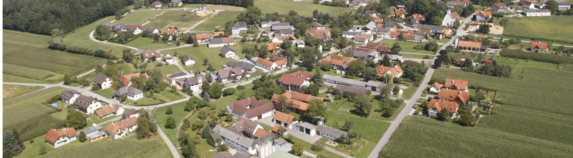 Luftaufnahme eines Dorfes mit mehreren Häusern, einige mit Solarpanelen auf ihren Dächern, umgeben von Bäumen und grünen Feldern.