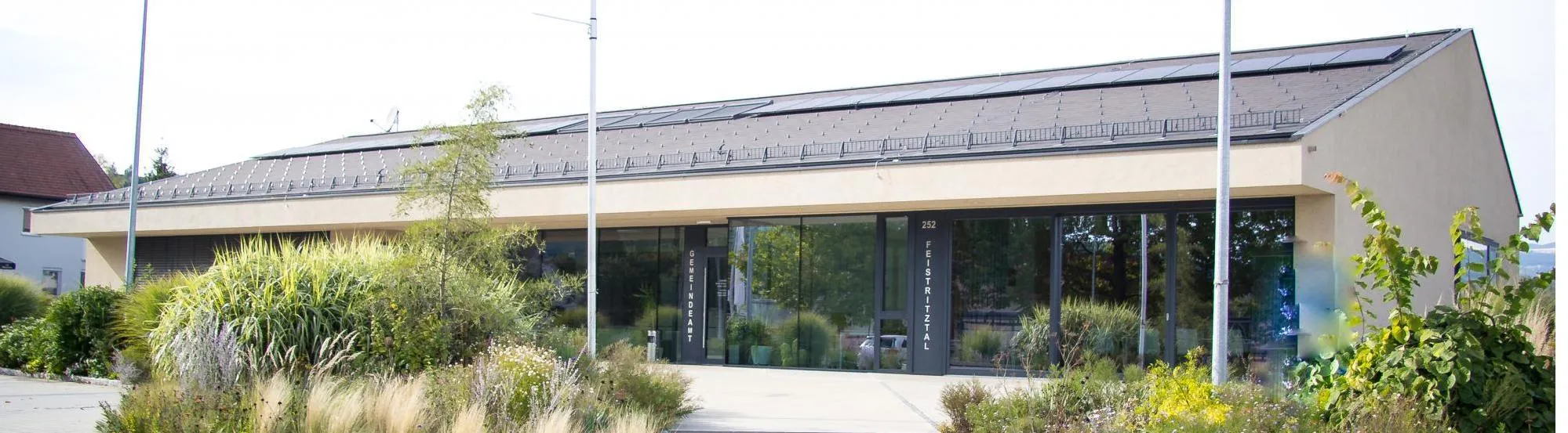 Ein Gebäude mit Solarpanels auf dem Dach hat einen Glaseingang und Pflanzen drumherum. Der Eingang hat den Text 'Gemeindeamt' links und 'Feistritzer' rechts.