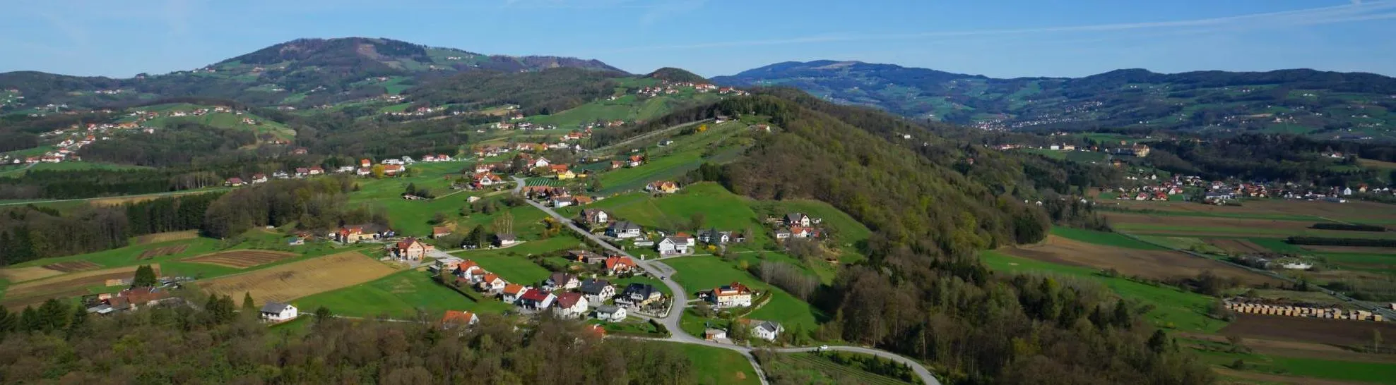 Luftaufnahme eines kleinen Dorfes in einer bergigen Region mit grünen Feldern, Häusern und kurvenreichen Straßen. Bäume und Büsche sind verstreut, und in der Ferne sind Berge unter einem klaren blauen Himmel sichtbar.