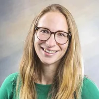 Eine Frau mit blondem Haar lächelt und schaut in die Kamera. Sie trägt eine Brille und ein grünes Oberteil. Sie scheint sich in einem Studio zu befinden.