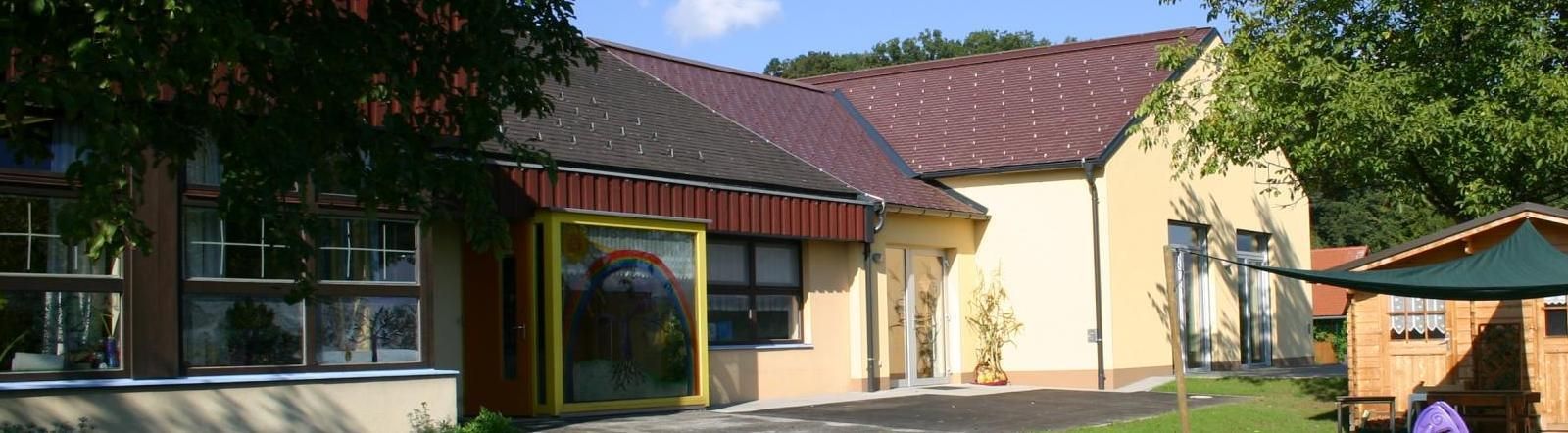 Ein Gebäude mit rotem Dach und gelben Wänden hat eine Glastür mit einem Regenbogen-Aufdruck. Eine Pflanze steht auf der rechten Seite.