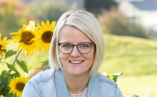 Eine Frau mit kurzen blonden Haaren und Brille lächelt vor Sonnenblumen.