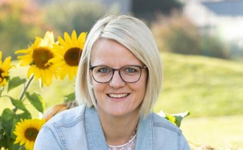 Eine Frau mit kurzen blonden Haaren und Brille lächelt vor Sonnenblumen.