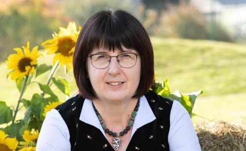 Eine Frau mit kurzen Haaren, die eine Brille und eine Halskette trägt, steht vor Sonnenblumen und einem grünen Feld.