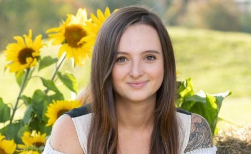 Eine Frau mit langen Haaren posiert für ein Foto in einem Feld mit Sonnenblumen. Sie lächelt fröhlich und hat ein Tattoo am Arm.