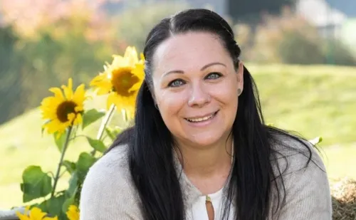 Eine Frau mit langen schwarzen Haaren lächelt vor Sonnenblumen. Sie trägt ein weißes Oberteil und einen grauen Cardigan. Hinter ihr sind verschwommenes grünes Gras und einige Sonnenblumen zu sehen.