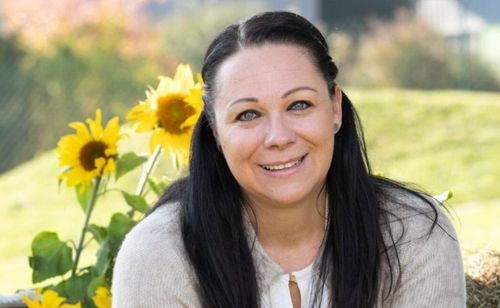 Eine Frau mit langen schwarzen Haaren lächelt vor Sonnenblumen. Sie trägt ein weißes Oberteil und einen grauen Cardigan. Hinter ihr sind verschwommenes grünes Gras und einige Sonnenblumen zu sehen.