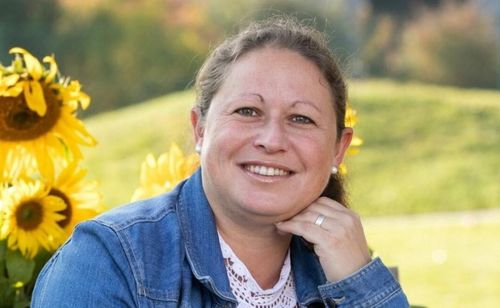Eine Frau lächelt und posiert für ein Foto mit einem Sonnenblumenfeld im Hintergrund.