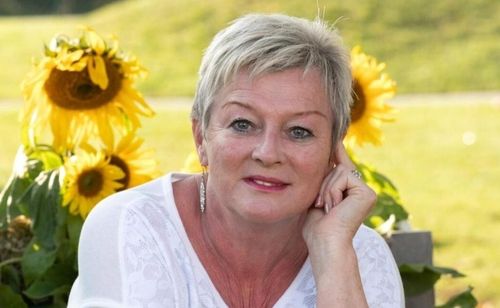 Eine ältere Frau mit kurzen blonden Haaren lächelt und posiert für ein Foto mit Sonnenblumen im Hintergrund. Sie trägt ein weißes Oberteil und Ohrringe.