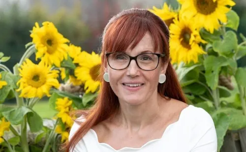 Eine Frau mit roten Haaren und Brille steht vor einem Feld mit Sonnenblumen.
