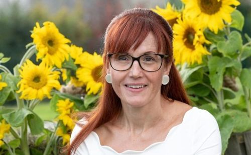 Eine Frau mit roten Haaren und Brille steht vor einem Feld mit Sonnenblumen.