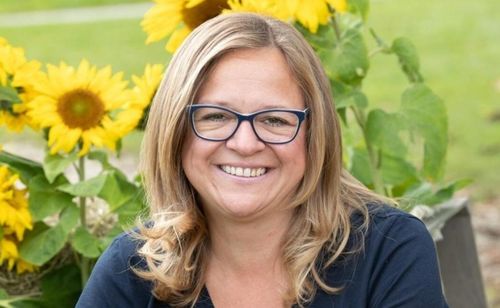 Eine Frau mit blonden Haaren und Brille lächelt vor einem Garten mit Sonnenblumen.