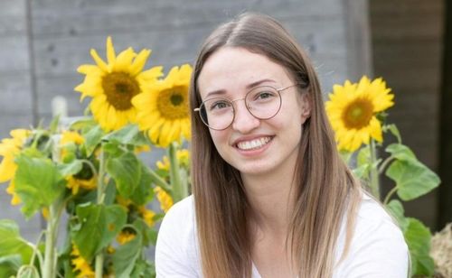 Eine lächelnde Frau mit langen Haaren und Brille steht vor einem Feld von Sonnenblumen.