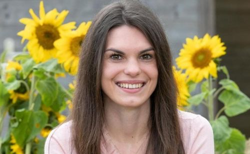 Eine Frau mit langen braunen Haaren lächelt vor einem Feld mit Sonnenblumen. Sie trägt ein rosa Oberteil und eine Halskette.