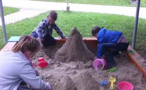 Vier Kinder spielen in einem Sandkasten, zwei mit Eimern und zwei mit Schaufeln, umgeben von einem Rasenbereich und einem Holzzaun.