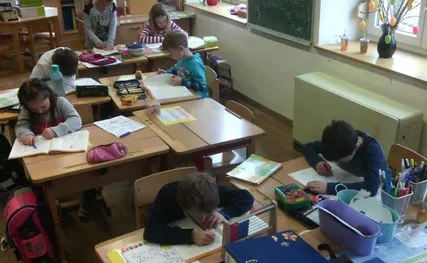 Kinder sitzen an Schreibtischen in einem Klassenzimmer, schreiben in ihre Notizbücher und arbeiten an verschiedenen Schulmaterialien. Auf den Tischen befinden sich Bücher, Stifte und andere Schulsachen. Eine Tafel ist an der Wand montiert.