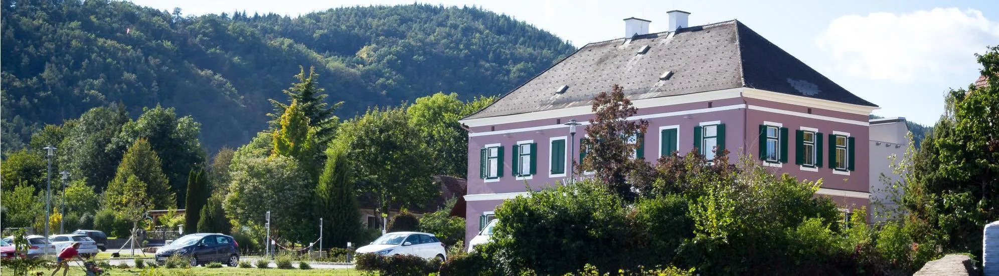 Ein rosafarbenes Haus mit grünen Fensterläden ist an einem sonnigen Tag zu sehen, umgeben von üppigen grünen Bäumen und Büschen. Zwei weiße Autos sind davor geparkt.