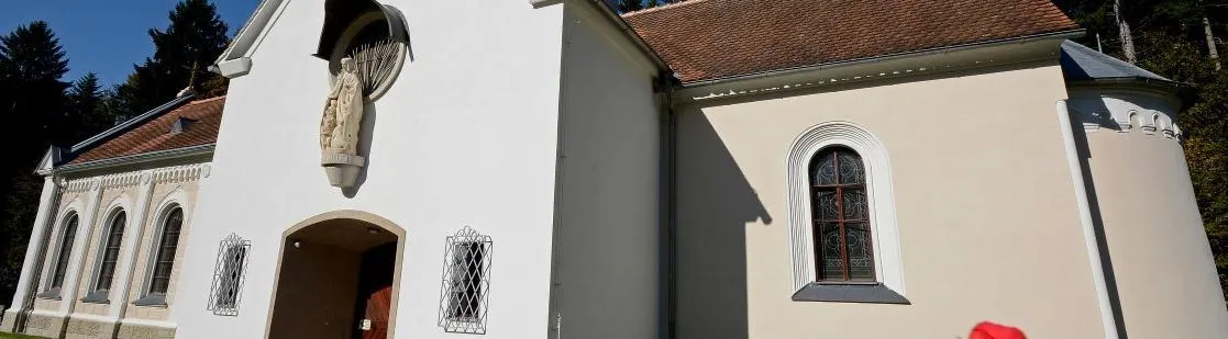 Eine weiße Kirche mit einer Statue über dem Eingang und einem Fenster mit kunstvollen Glasdesigns. Das Gebäude hat ein braunes Dach und Metallgitter an den Fenstern.