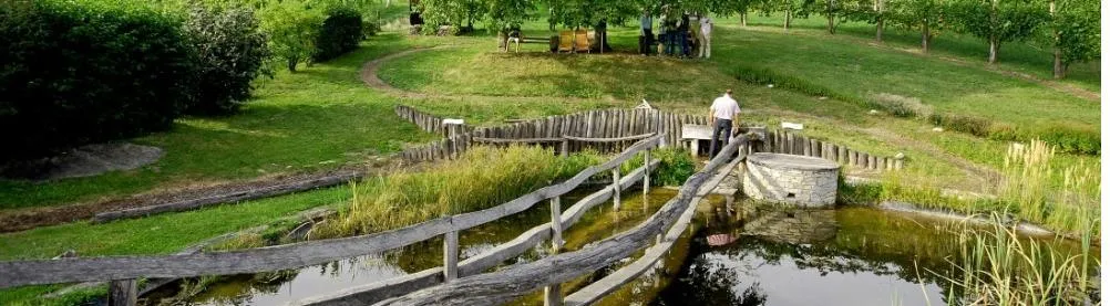 Eine Luftaufnahme eines Parks mit einem Holzzaun und einem Mann, der darauf steht. Hinter dem Zaun befindet sich ein kleiner Teich mit Wasser. Es gibt einen Rasenbereich mit einer Holzbank und einige Personen, die in der Nähe stehen.