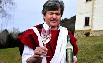 Ein Mann in einem roten und weißen Gewand hält ein Weinglas mit Weißwein. Er steht vor einem Gebäude mit einer Steinmauer, möglicherweise einem Weingut.