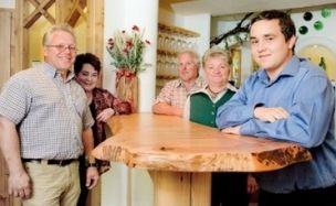 Fünf Personen stehen hinter einem hölzernen Tresen und posieren für ein Foto. Der Tresen hat eine natürliche Holzstruktur. Sie befinden sich in einem Raum mit einer hölzernen Wand und einer Blumenvase auf einem Tisch.