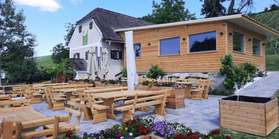 Eine hölzerne Außengastronomie mit Bänken und Tischen, umgeben von Blumen und Pflanzen. Ein Haus mit braunem Dach und grünem Schild im Hintergrund.