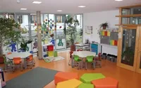 Ein Innenraum ähnelt einem Klassenzimmer mit Glasfenstern, ausgestattet mit runden Tischen und Stühlen. Topfpflanzen schmücken den Raum, und ein Teppich bedeckt den Boden. Bunte Kissen sind auf dem Boden verstreut.