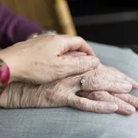 Die Hände einer älteren Person werden von einer anderen Person gehalten. Die ältere Frau trägt einen Ring und hat Falten an ihren Händen.