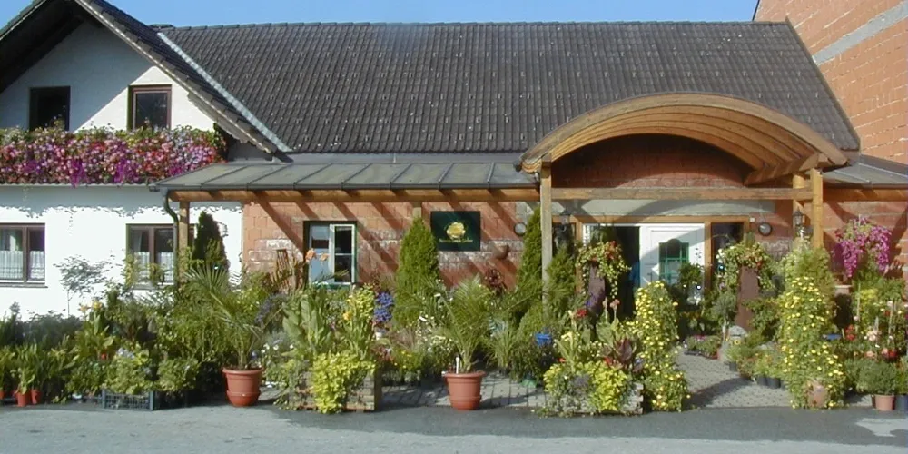 Ein Haus mit einem geneigten Dach und einem mit Blumen geschmückten Balkon steht vor einem Garten mit Topfpflanzen und Blumen. Eine hölzerne Pergola und ein grünes Schild sind in der Nähe.