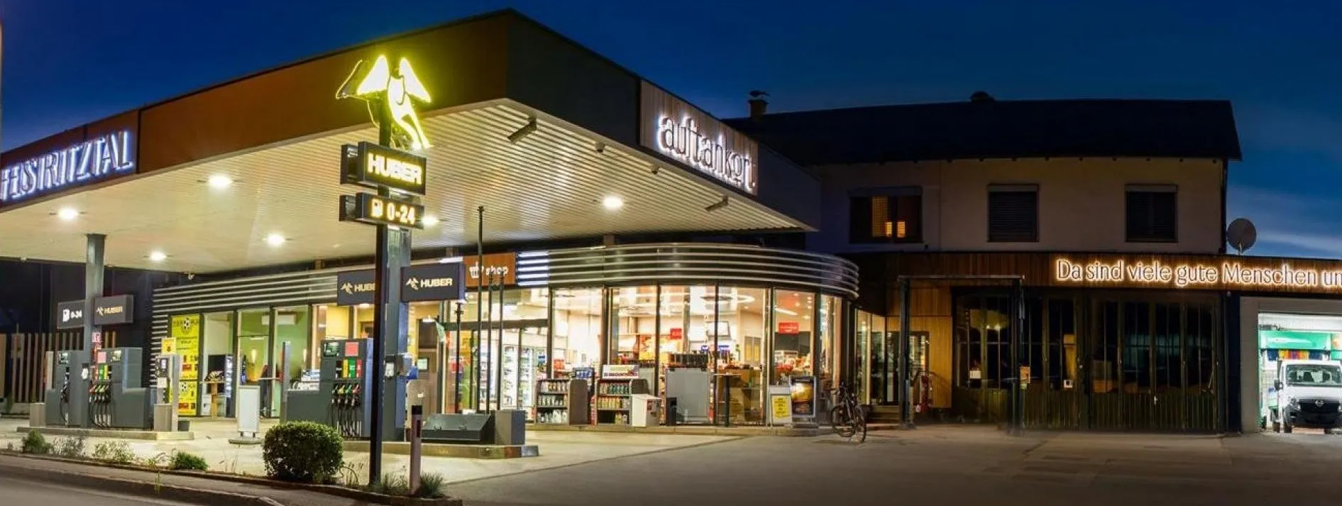 Ein beleuchteter Auftanker-Shop bei Nacht, mit Produkten im Schaufenster, einer Tankstelle und einem geparkten Fahrrad davor.