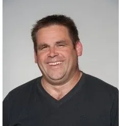 Ein lächelnder Mann mit kurzen Haaren, der ein schwarzes V-Neck-Shirt trägt. Er schaut direkt in die Kamera.