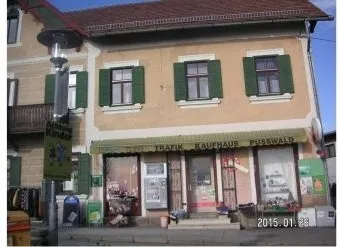 Ein zweistöckiges Gebäude mit grünen Fensterläden und einem grünen Vordach, beschriftet mit 'TRAFEK KAUFHAUS PUSSWALD'. Das Gebäude hat mehrere Fenster und einen Balkon. Auf dem Bürgersteig befinden sich verschiedene Gegenstände, einschließlich eines Mülleimers und einer Straßenlaterne. Das Bild ist vom 28.01.2015.