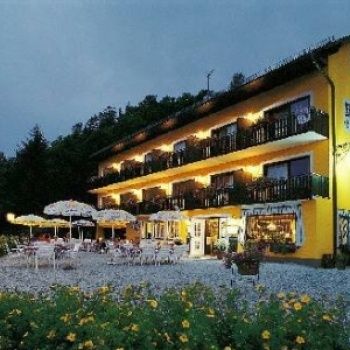 Ein gelbes Hotel mit mehreren Balkonen und Fenstern, nachts beleuchtet. Ein Außenbereich mit Tischen, Stühlen und Sonnenschirmen. Vor dem Hotel stehen Pflanzen und Blumen.