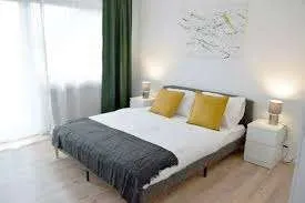 Ein ordentlich eingerichtetes Schlafzimmer mit weißen Wänden, Holzböden und einem großen Bett. Zwei Nachttische mit Lampen flankieren das Bett, und ein weißer Vorhang bedeckt ein Fenster.