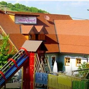 Ein Haus mit einem braunen Dach und einem Schild, das Die Morcherl Cafe liest. Vor dem Haus steht ein bunter Spielplatz und ein Zaun. Draußen steht ein Tisch und Stühle.