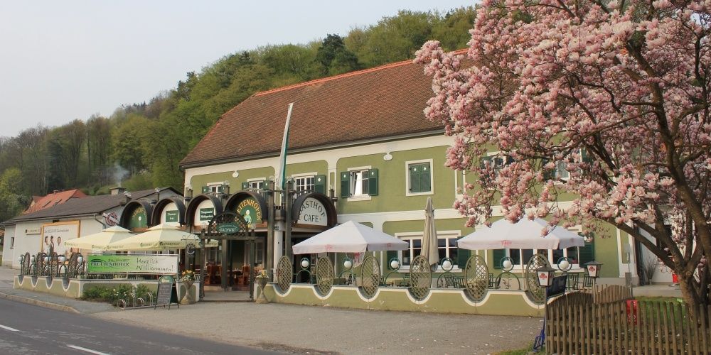 Ein grünes Gebäude mit dem Namen 'Gasthof', umgeben von Tischen und Sonnenschirmen. Ein Baum mit rosa Blüten davor.