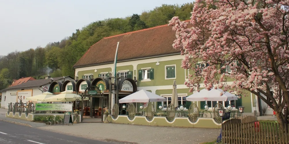 Ein grünes Gebäude mit dem Namen 'Gasthof', umgeben von Tischen und Sonnenschirmen. Ein Baum mit rosa Blüten davor.
